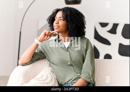 Eine nachdenkliche afroamerikanische Frau, die in entspannter Atmosphäre auf einer Couch sitzt und durch die in Träumen verlorenen Fenster blickt, verbringt eine ruhige, charmante Dame ihre Freizeit zu Hause Stockfoto