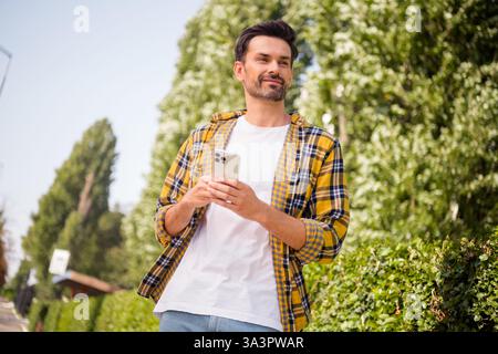 Lässig gekleideter junger Mann, der ein Smartphone hält, während er einen angenehmen Spaziergang im Stadtpark unter dem Sommerlicht genießt Stockfoto