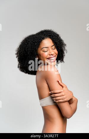 Eine ausgelassene afroamerikanische Frau mit einem breiten Lächeln steht mit geschlossenen Augen, verkörpert Freude und unbeschwerten Geist, lockiges Haar ergänzt perfekt die neutrale Kleidung vor hellem Hintergrund Stockfoto