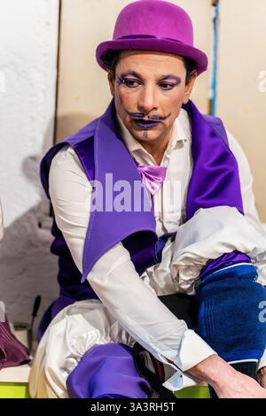 Clown in bunten Kostümen und Make-up macht sich hinter der Bühne bereit, passt Kleidung an und blickt mit fokussiertem Ausdruck nach unten Stockfoto