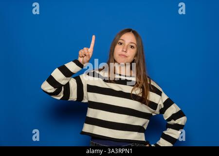 Hispanic Teenager Mädchen ca. 15 Jahre mit gestreiftem Pullover über blauem Hintergrund, der den Finger nach oben zeigt und von einem brillanten Gedanken inspiriert aussieht Stockfoto