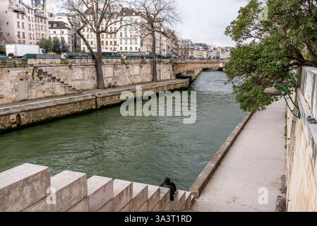 Das Ufer der seine an einem bewölkten Tag. Eine nicht identifizierte Person, die am Ufer mit einem Handy sitzt Stockfoto
