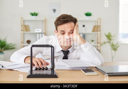Mann, der am Schreibtisch sitzt, am Arbeitsplatz im Büro, mit Newtons Wiege Balancebälle, die von der Arbeit müde sind Stockfoto