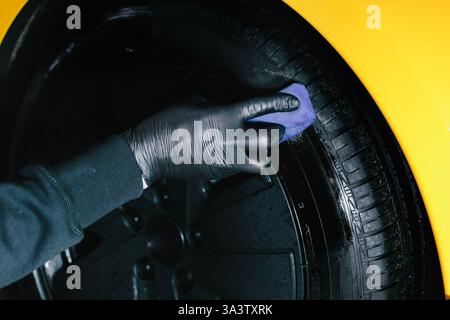 Der Mechaniker wendet ein Produkt mit einem blauen Applikator auf einen schwarzen Reifen an. Schwärzung des Reifenrads des Autos mit Schwamm mit chemischer Schwärzung des Gummis in Stockfoto