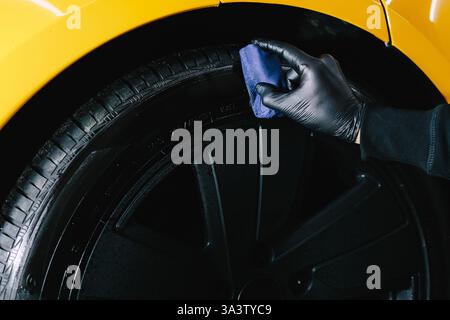 Der Mechaniker wendet ein Produkt mit einem blauen Applikator auf einen schwarzen Reifen an. Schwärzung des Reifenrads des Autos mit Schwamm mit chemischer Schwärzung des Gummis in Stockfoto