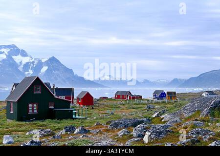 Ein malerischer Blick auf das unbewohnte Dorf Qoornoq mit farbenfrohen traditionellen Inuit-Häusern am felsigen Fjordufer und schneebedeckten Bergen im Hintergrund Stockfoto