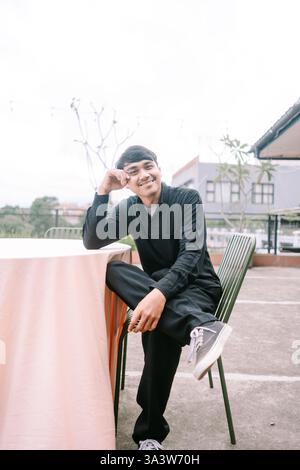 Ein junger asiatischer Mann sitzt und entspannt sich draußen. Er ist lässig in schwarzer Kleidung und grauen Schuhen gekleidet und liegt bequem auf einem Stuhl. Er hat eine entspannte, Stockfoto