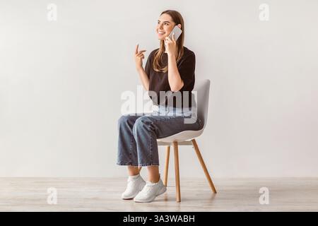 Die brünette Frau sitzt bequem im Stuhl, während sie mit dem Smartphone spricht Stockfoto