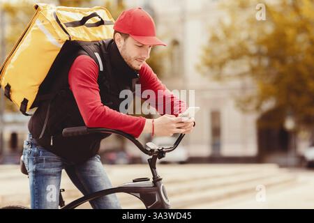 Delivery Guy In Uniform Mit Handy Sitzen Auf Fahrrad Im Freien Stockfoto