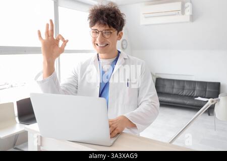 Männlicher medizinischer Praktikant mit Laptop zeigt OK an der Rezeption in der Klinik Stockfoto