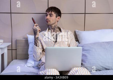 Junger Mann im Schlafanzug mit elektronischer Einwegzigarette mit Laptop auf dem Bett zu Hause Stockfoto