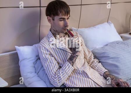 Junger gutaussehender Mann im Pyjama, der elektronische Einweg-Zigarette auf dem Bett zu Hause raucht Stockfoto