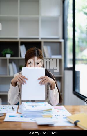 Junge Frau in beigefarbenem Langarmhemd, verspielt ihr Gesicht mit einem leeren Laken an einem Schreibtisch, überfüllt mit Papierkram und Dokumenten. Stockfoto