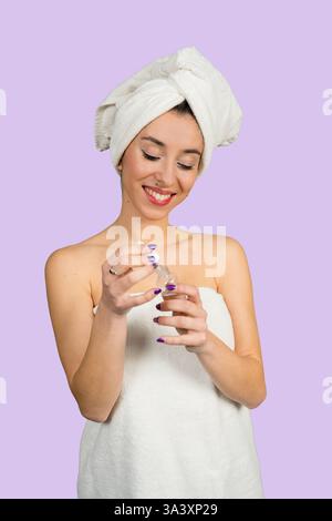 Die schöne junge Frau lächelt, während sie nach dem Duschen Serum auf ihr Gesicht aufträgt und ein Handtuch trägt Stockfoto