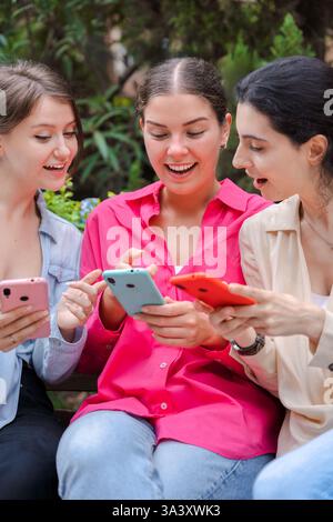 Junge Frauen sitzen auf einer Bank, lächeln und benutzen Smartphones in einem Park im Freien Stockfoto