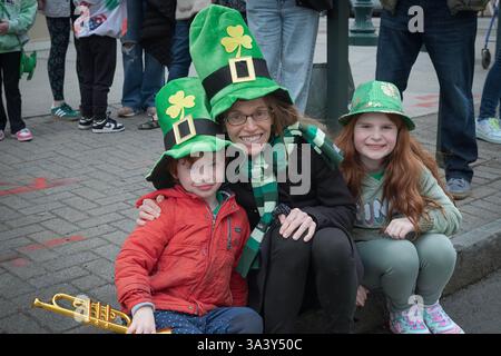Eine fröhliche Familie, die 2025 die Peekskill Saint Patrick's Day Parade in Westchester, NY, ansieht, während sie auf einem Bordstein sitzt Stockfoto