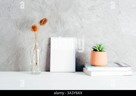 Minimalistisches Innendekor mit einem leeren weißen Rahmen, getrockneten Blumen in einer Glasvase und einem Sukkulenten auf Büchern vor einer strukturierten grauen Wand. Modell Stockfoto
