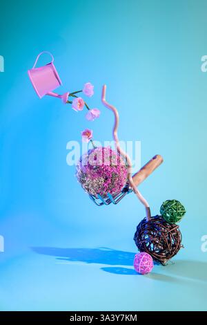 Die pinke Gießkanne gießt Wasser auf wachsende Blumen, die von einem Handrechen gehalten werden, der auf dekorativen Korbbällen in einem surrealen Frühlingskonzept mit Licht ausgeglichen ist Stockfoto