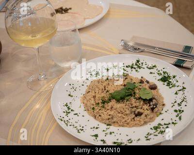 Pilzrisotto, serviert auf einem weißen Teller und garniert mit Petersilie, begleitet von einem Glas Weißwein und einem Glas Wasser, auf einem Restauranttisch Stockfoto