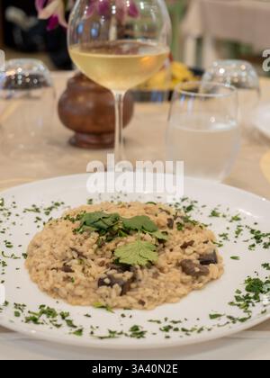 Köstliches Pilzrisotto garniert mit Petersilie und Koriander serviert auf einem weißen Teller in einem eleganten Restaurant mit einem Glas Weißwein und einem Glas Stockfoto