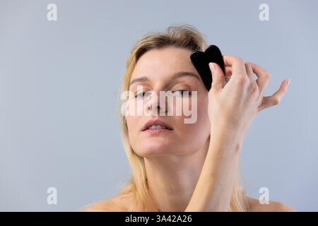 Das Bild zeigt eine Frau, die eine Gesichtsmassage mit einem schwarzen Gua-Sha-Werkzeug durchführt, um Hautpflege und Wohlbefinden zu fördern und gleichzeitig ihre na zu zeigen Stockfoto
