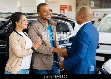 Afroamerikanisches Paar unterzeichnet im Autohändler eine Vereinbarung über den Kauf von Fahrzeugen und den Handschlag mit dem Verkäufer Stockfoto