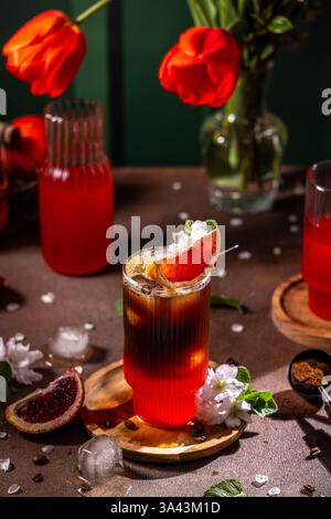 Das trendige Rot-Orange- und Kaffeegetränk wird in einem stilvollen Glas mit Eis serviert, garniert mit einem Stück roter Orange und zarten weißen Blüten Stockfoto