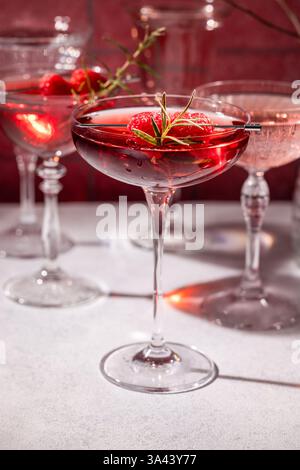 Coupé-Gläser gefüllt mit leuchtend roten Cocktails, verziert mit frischen Himbeeren und einem duftenden Rosmarinzweig, schaffen eine raffinierte und festliche Atmosphäre Stockfoto