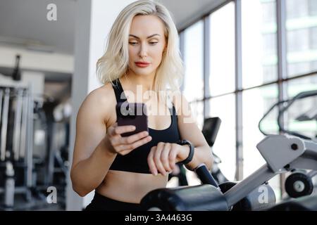 Blonde Athletin, die Fitness-App auf dem Smartphone nutzt und Smartwatch während des Workouts im Fitnessstudio überprüft Stockfoto