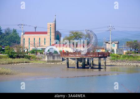 Gwangyang City, Südkorea - 3. Oktober 2021: Eine große Metallfischskulptur steht auf einer hölzernen Plattform über dem Seomjin River, mit einer Kirche im B Stockfoto