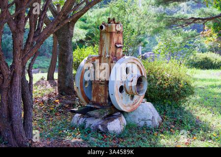 Gwangyang, Südkorea - 3. Oktober 2021: Eine verlassene und verrostete Industriemaschine sitzt auf einem Steinfuß unter dem Schatten von Bäumen und verschmilzt in Stockfoto