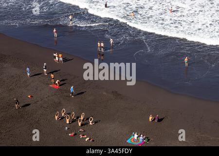 Ein lebhafter Blick aus der Luft auf einen schwarzen Sandstrand, wo die Menschen verschiedene Aktivitäten unter der Sonne genießen. Einige Menschen waten in den Wellen des Ozeans Stockfoto