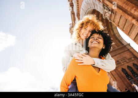 Zwei glückliche Frauen genießen an einem sonnigen Tag einen Huckepack-Ritt unter dem Arc de triomf in barcelona, spanien Stockfoto