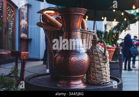 Vintage-Kupferkrug, geflochtene Weinflasche und französisches Baguette in Korb, der auf Holzfässern steht. Antike Metallflasche auf Holztisch am Bürgersteig Stockfoto