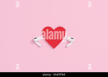 Ansicht von oben auf weiße Ohrhörer und rotes Papierherz auf pastellrosa Hintergrund. Mockup Vorlage für Valentinstag. Liebe und Technologiekonzept Stockfoto