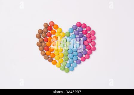 Bunte Süßigkeiten bilden eine Herzform auf weißem Hintergrund. Regenbogenfarbene Süßigkeiten. Glücklichkeits- und Fröhlichkeits-Konzept, Blick von oben Stockfoto