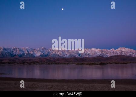 Blick am frühen Morgen auf die schneebedeckte Sierra Nevada mit dem Mond darüber, der sich im Wasser von neuem Regen spiegelt. Stockfoto