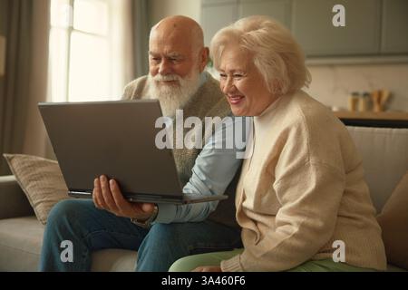 Fröhliches Seniorenpaar, das Laptop benutzt, während es auf dem Sofa im Wohnzimmer sitzt Stockfoto