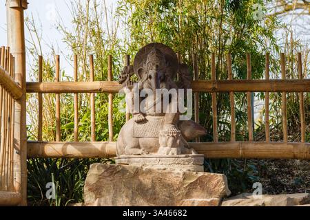 Stone Ganesha Statue im Garten. Eine Steinstatue der hinduistischen Gottheit Ganesha, die auf einem Felsen sitzt, umgeben von Bambuszäunen und Grün. Stockfoto