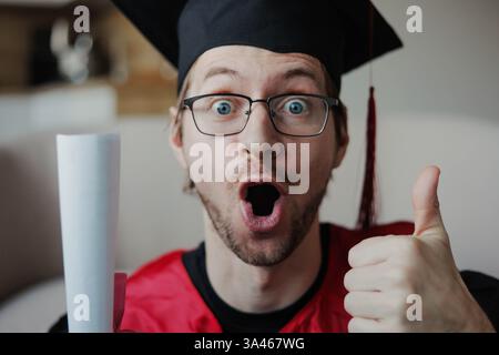 Aufgeregter, lustiger Mann in schwarzem Abschlusskleid und Mütze mit Diplom. Nahaufnahme. Stockfoto