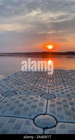 Ein schwimmendes Dock mit strukturierter Oberfläche erstreckt sich bei Sonnenuntergang über einen stillen See und reflektiert die warmen Farben des Himmels auf dem Wasser. Fotografiert in einer Natur Stockfoto