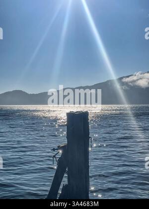 Sonnenlicht fließt in Strahlen über ein ruhiges Gewässer und erzeugt glitzernde Reflexionen um einen alten Holzpfosten mit weit entfernten Bergen im Rücken Stockfoto