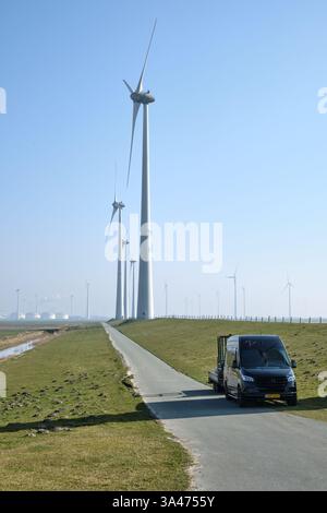 10. März 2025: Eemshaven, Niederlande: Windräder entlang einer Landstraße mit einem schwarzen Firmenwagen im Vordergrund in der Nähe einer Energieanlage. Stockfoto