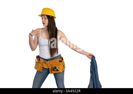 Eine junge Bauarbeiterin mit Schutzhelm und Werkzeuggürtel macht eine Pause und wischt ihr Gesicht ab, während sie ihr Jeansshirt auszieht. Sie drückt Exhau aus Stockfoto