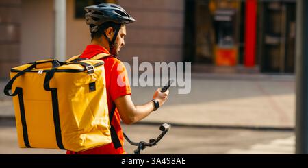 Junge männliche Radfahrer mit Kurier Liefertasche mit Handy auf der Straße, um Adresse des Kunden in der mobilen Anwendung zu suchen Stockfoto