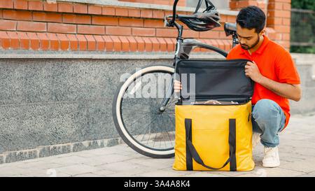 Verpackung der Bestellung in Thermobeutel. Serious Kerl Kurier mit Bart in Uniform mit Fahrrad sieht an gelbe Tasche Stockfoto