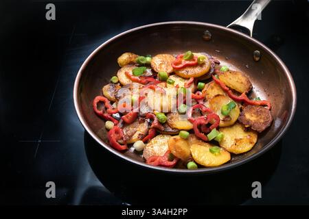 Knusprige gebratene Kartoffeln, rote Paprika und grüne Frühlingszwiebeln in einer Pfanne auf einem schwarzen Kochfeld in der Küche zu Hause, kochen mit Gemüse und Gemüse Stockfoto