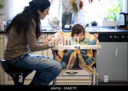 Eine Mutter sitzt auf einem hohen Stuhl und füttert ihr Kleinkind, das auf einem Hochstuhl sitzt. Das Duo genießt zusammen in der Küche die Mahlzeiten. Stockfoto