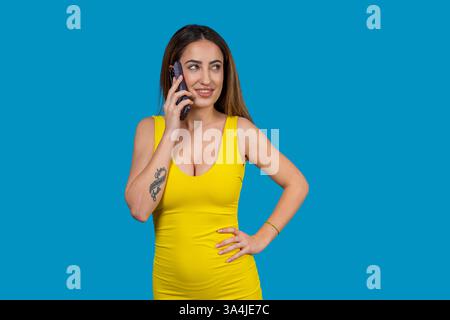 Schwangere Frau in gelbem Kleid mit Handy, lächelt und blickt weg auf blauem Hintergrund Stockfoto
