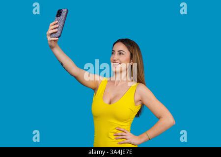 Junge Frau, die mit ihrem Smartphone ein Selfie macht, lächelt und posiert auf blauem Hintergrund Stockfoto